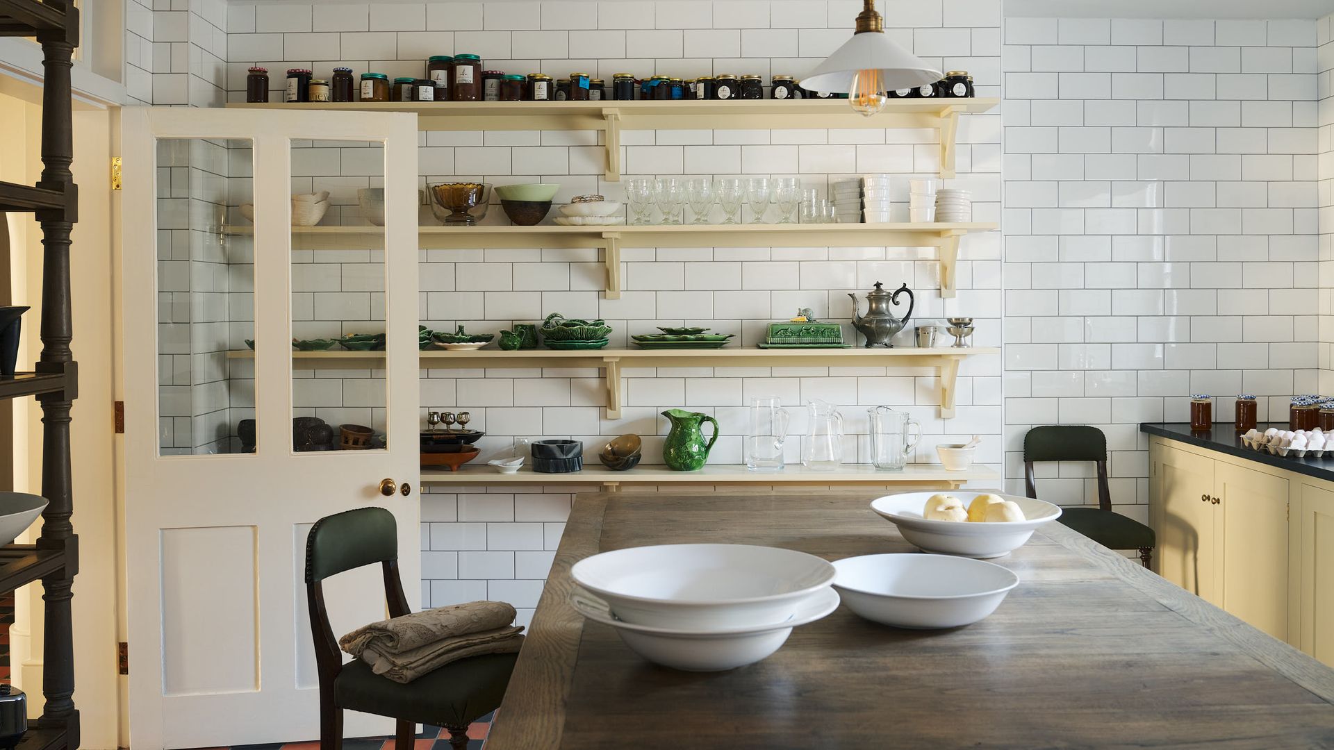 Cocina con isla de madera, pared de azulejos blancos brillantes, baldas de color vainilla con botes de mermelada, cristalería y vajilla, taburete alto de madera