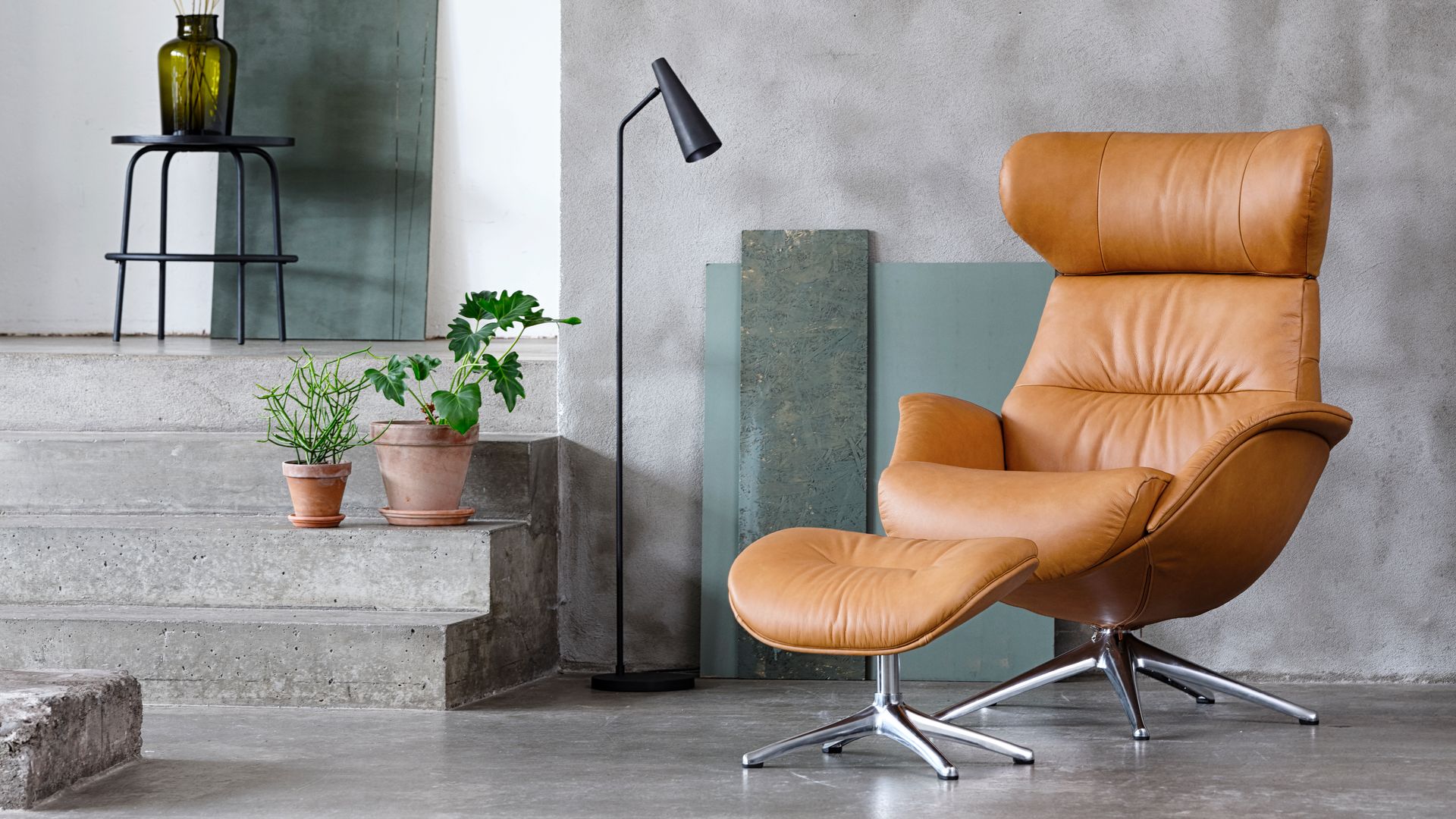 Butaca de cuero marrón con reposapiés a juego, suelo y paredes grises, lámpara de pie, macetas en el suelo