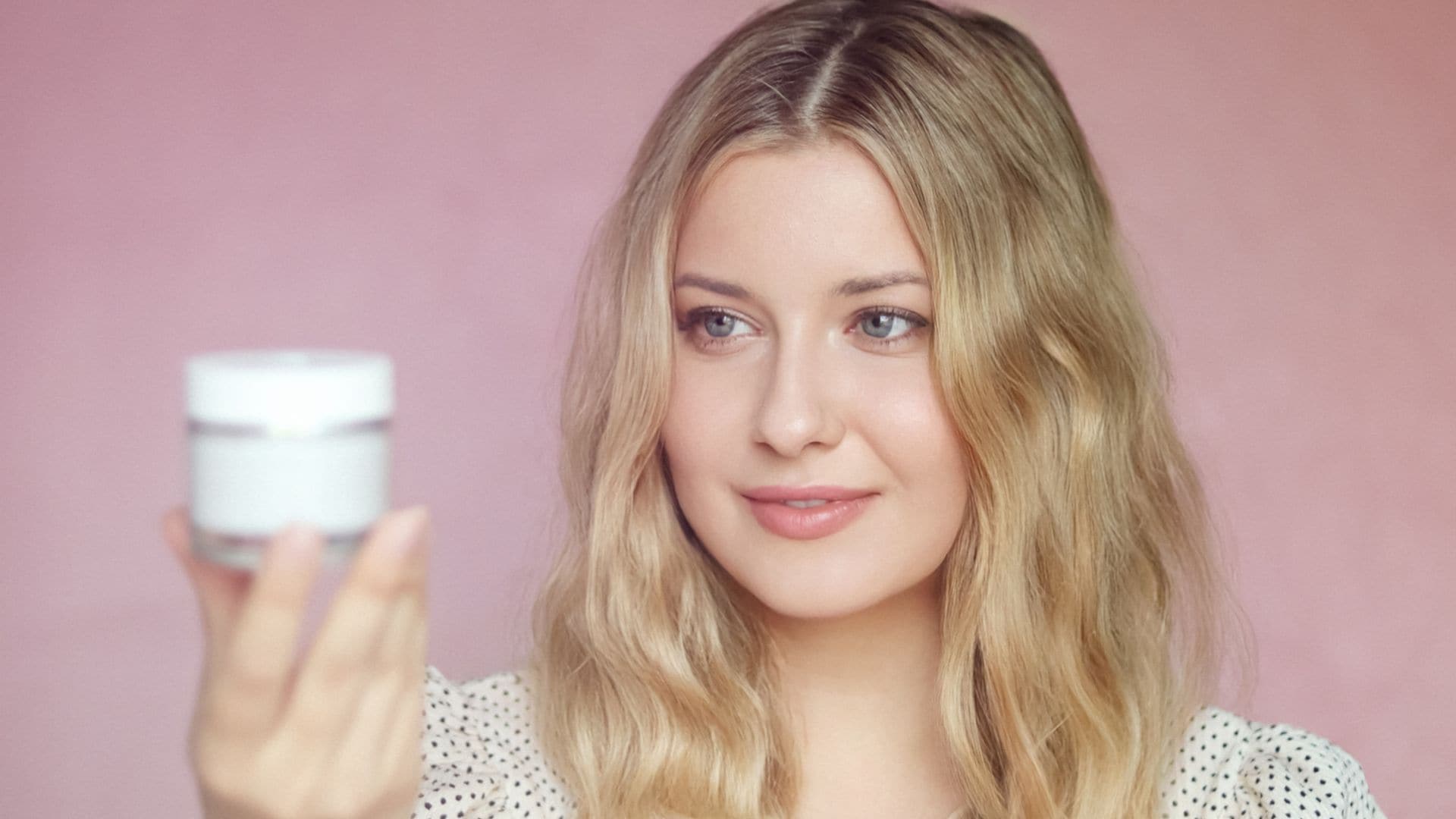 Mujer rubia joven sosteniendo una crema en su mano a la altura del rostro