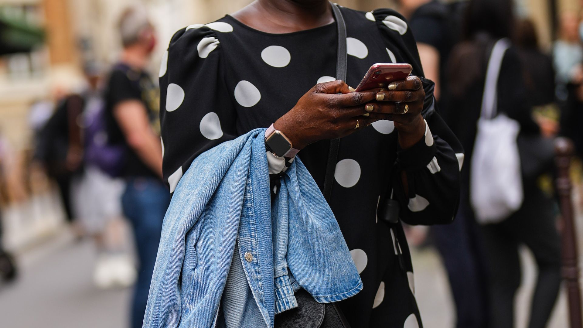Street Style At  Paris Fashion Week - Menswear Spring/Summer 2022. Mujer tecleando en un teléfono móvil con un reloj inteligente. Va con un vestido de lunares en blanco y negro, además de una cazadora vaquera en el brazo.