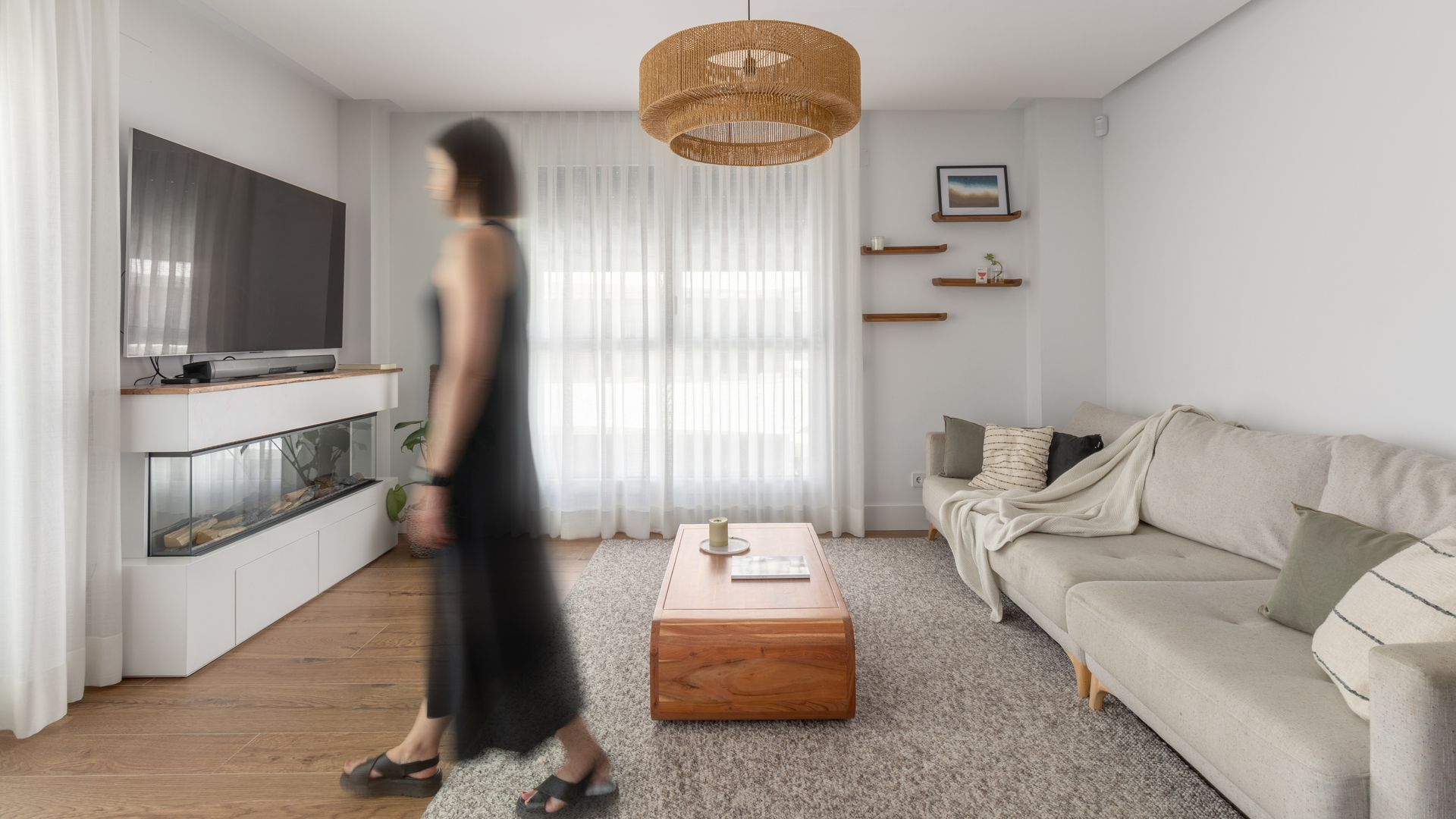 Salón en blanco con mujer pasando, sofá blanco, mueble de televisión con chimenea debajo, alfombra, cortinas, mesa de centro de madera