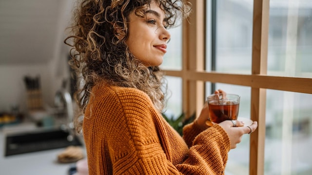 young woman holding a cup of tea and standing by the window tea time rituals 