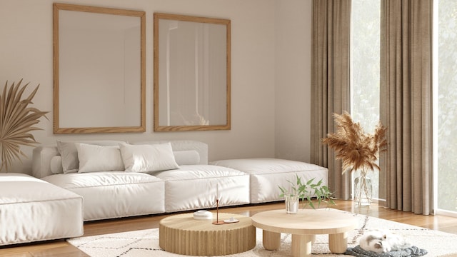 Salón decorado en estilo 'boho', en tonos blancos y madera clara, además de beige. Sofá cama chaise longue al fondo, con espejos en la pared, una alfombra geométrica en el centro y dos mesas bajas.