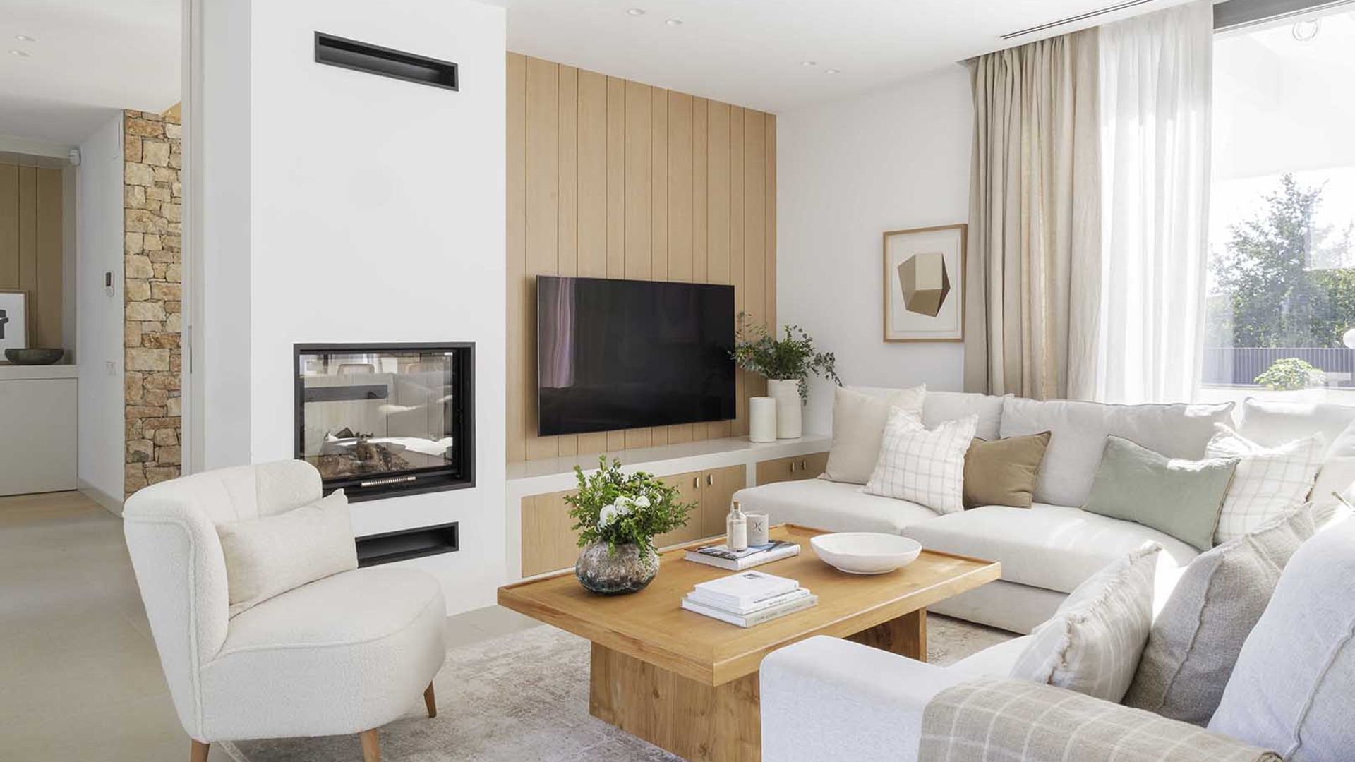 Salón con chimenea con sofá blanco, mesa de centro de madera, alfombra, cortinas y visillos, televisión colgada pared