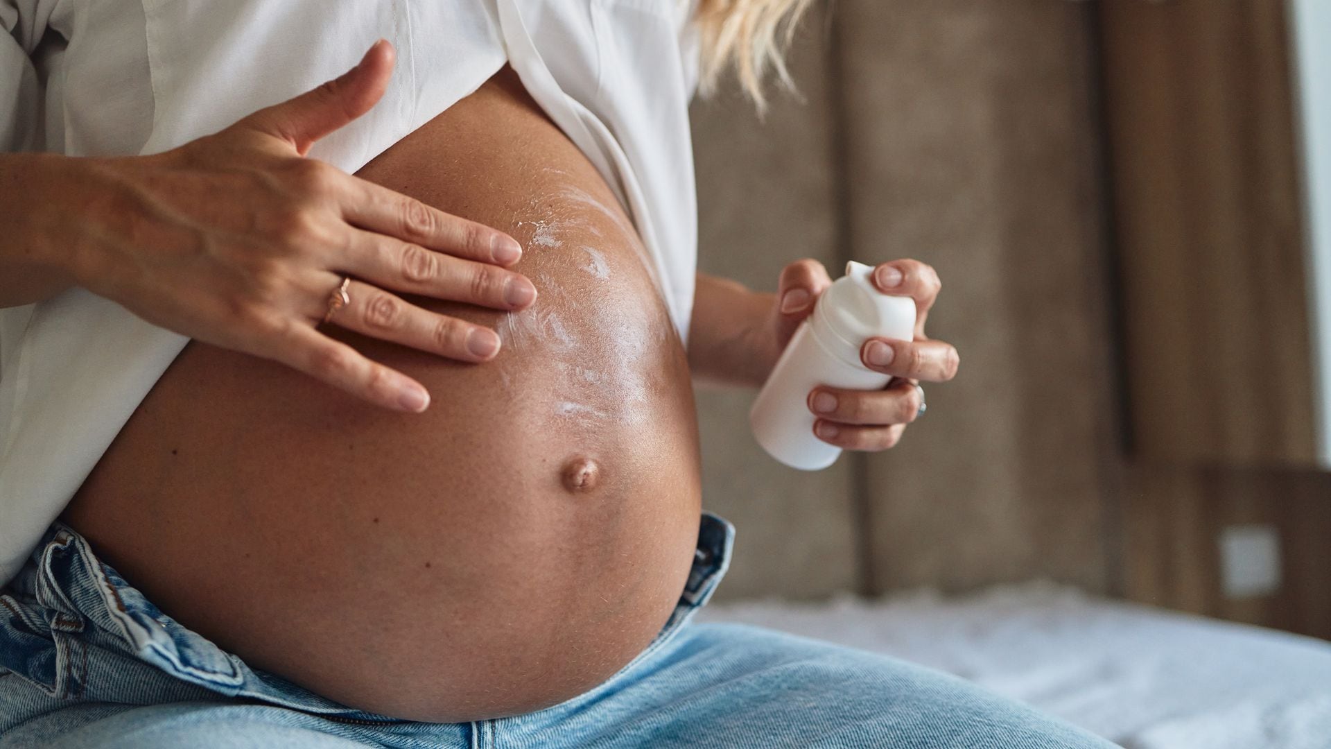 Mujer embarazada aplicándose crema en la barriga, con unos pantalones vaqueros y una camisa blanca. Plano detalle.
