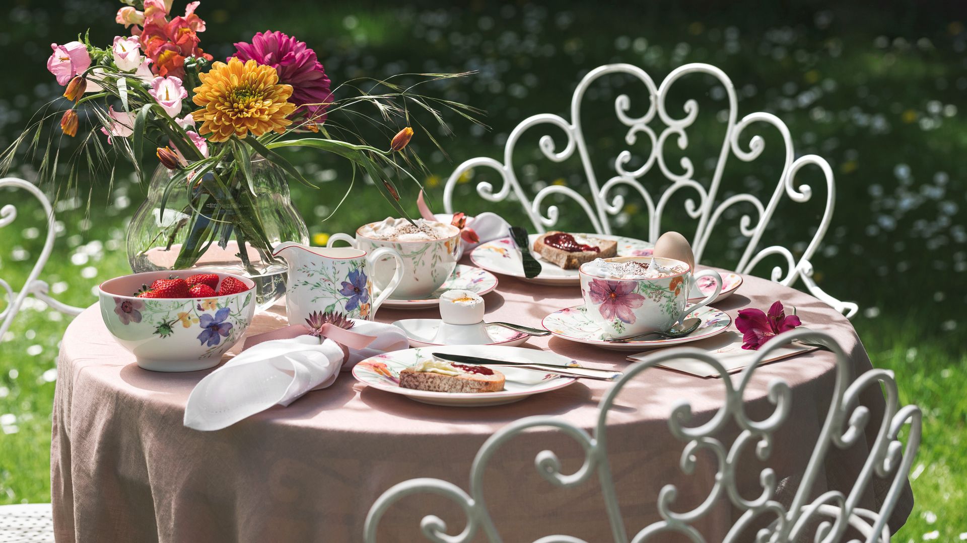 Mesa en el jardín con mantel rosa, sillas de hierro blanco, vajilla de flores, jarrón con flores y servilletas blancas