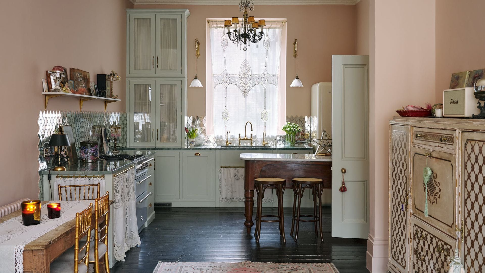 Cocina con mesa de madera con el sobre de mármol, lámpara de techo sillas de bambú, paredes de color rosa palo, apliques de pared dorados, velas