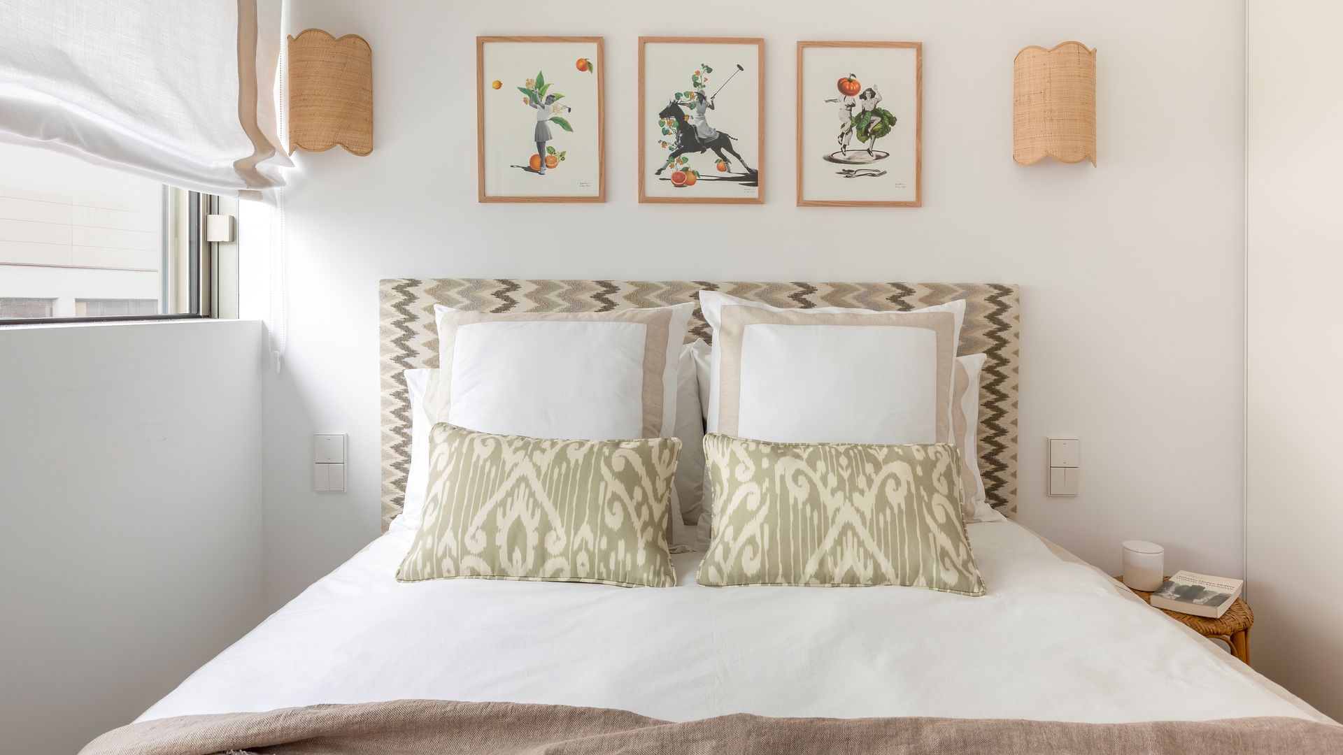 Dormitorio con cabecero tapizado, estor blanco, ropa de cama blanca, cojines, apliques de pared de fibras vegetales, cuadros sobre el cabecero 