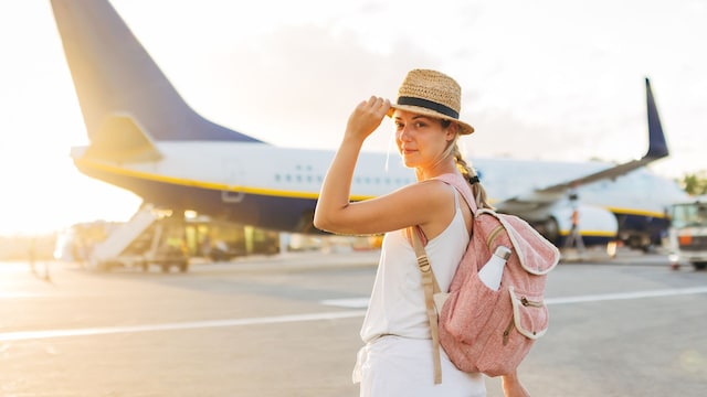 Mujer sonriendo a la cámara con la mano en el sombrero en la pista de un aeropuerto con una mochila en la espalda y una maleta de mano, con un avión al fondo.
