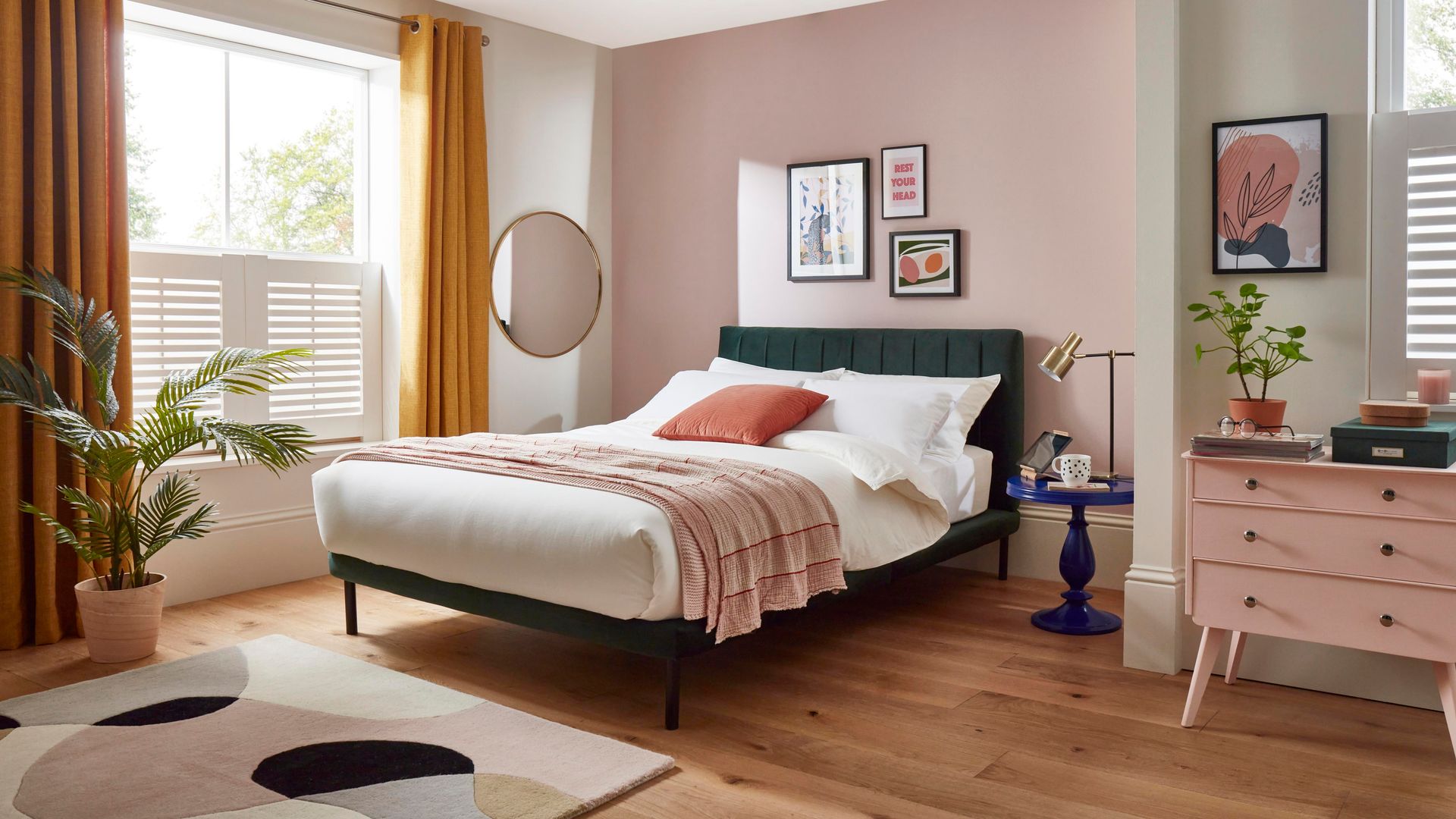 Dormitorio con paredes rosas, gran ventanal con cortinas color mostaza, suelo de madera, alfombra, cabecero tapizado en verde, cuadros, cómoda y plantas