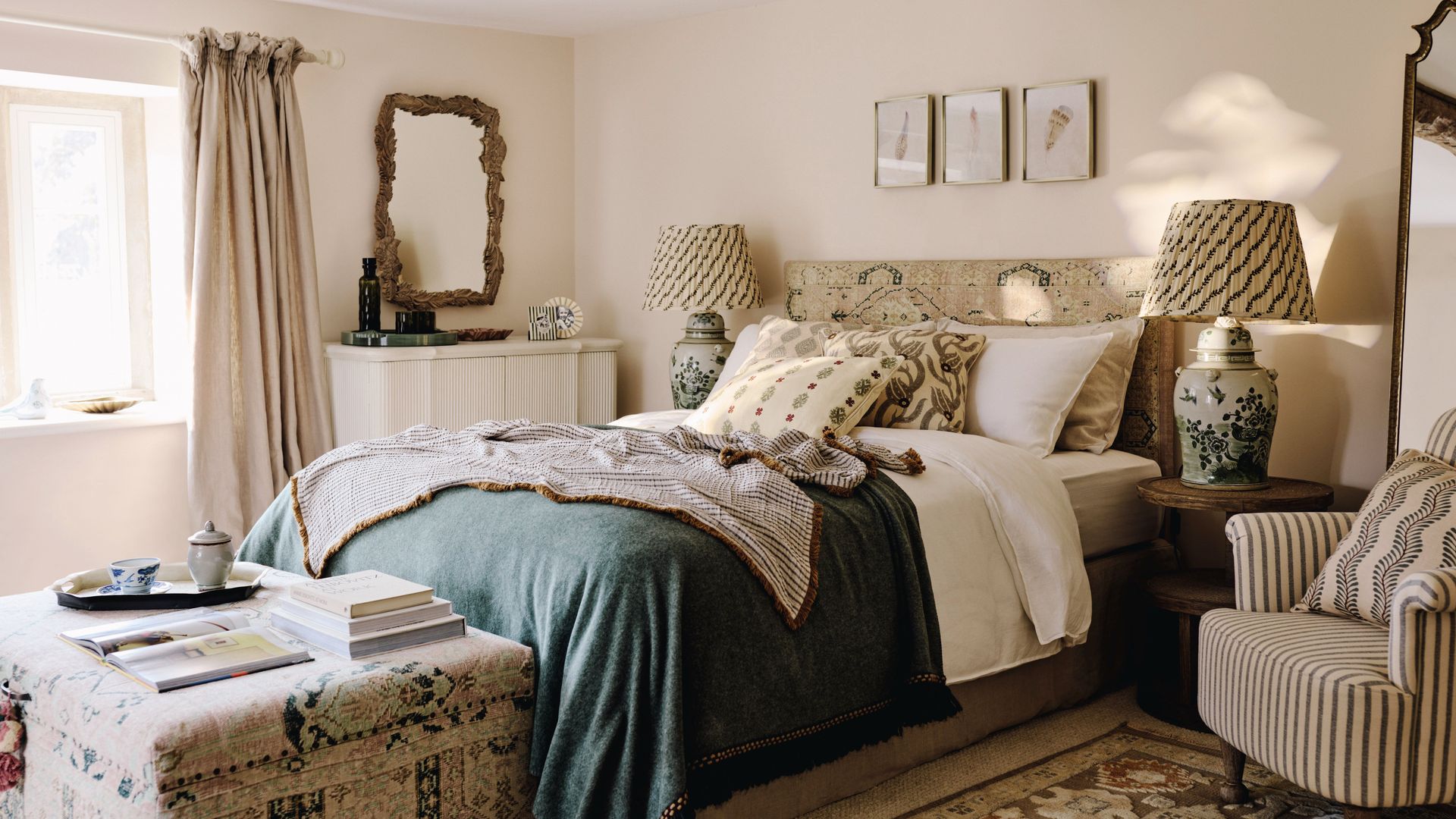 Dormitorio de estilo cottage con cabecero tapizado, banqueta a los pies, lámparas de sobremesa con la pantalla de tela, butaca, viga de madera en el techo 