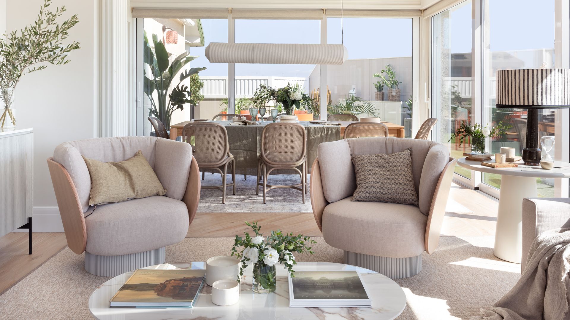 Salón comedor con salida a la terraza, decorado en tonos beiges y arenas, butacas tapizadas, sillas de comedor de rejilla, vigas de madera blanca, alfombras, lámpara de techo 