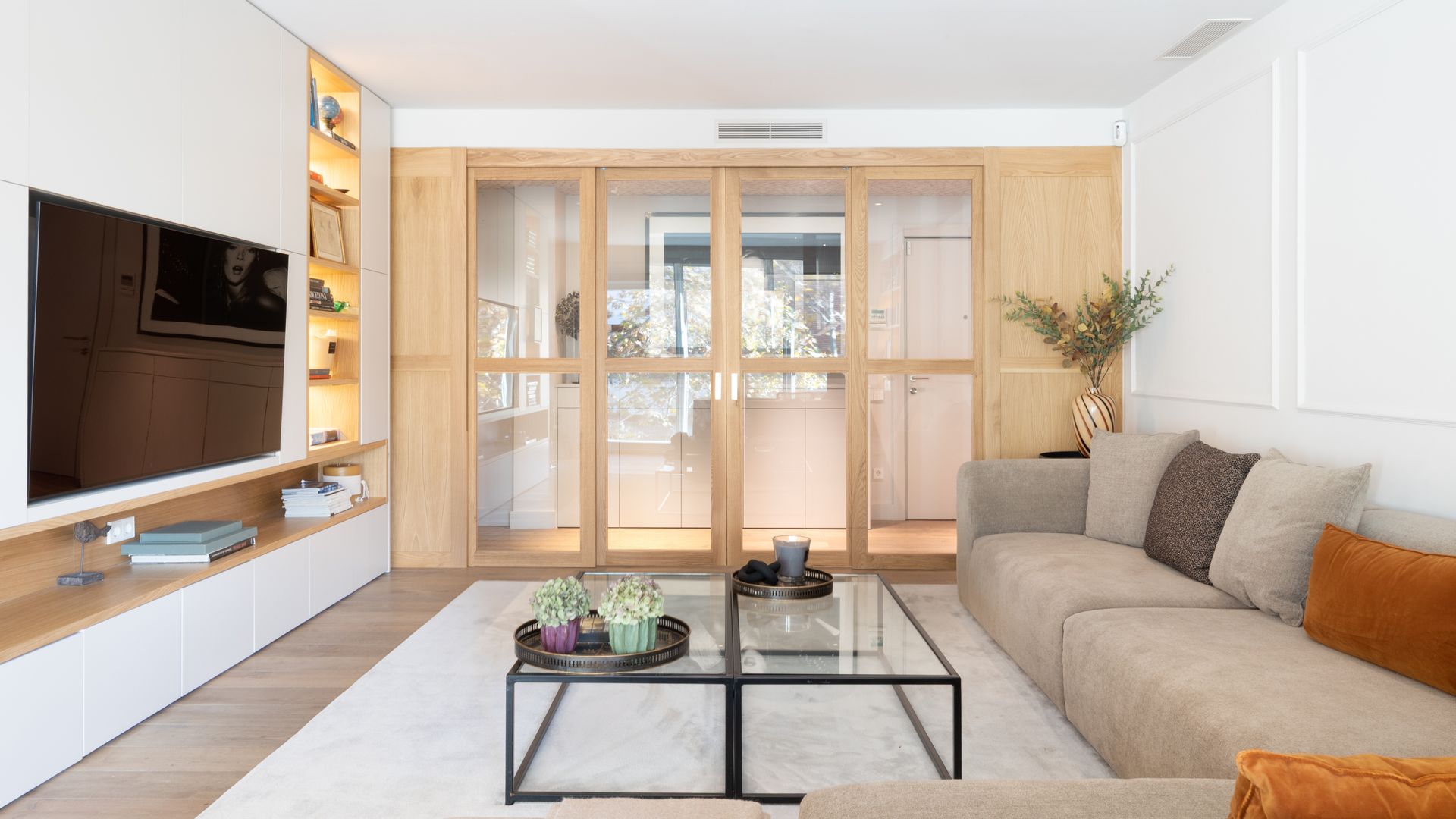 Salón con sofá beige, mueble de televisión a medida, molduras en el pared del sofá, mesa de centro de cristal