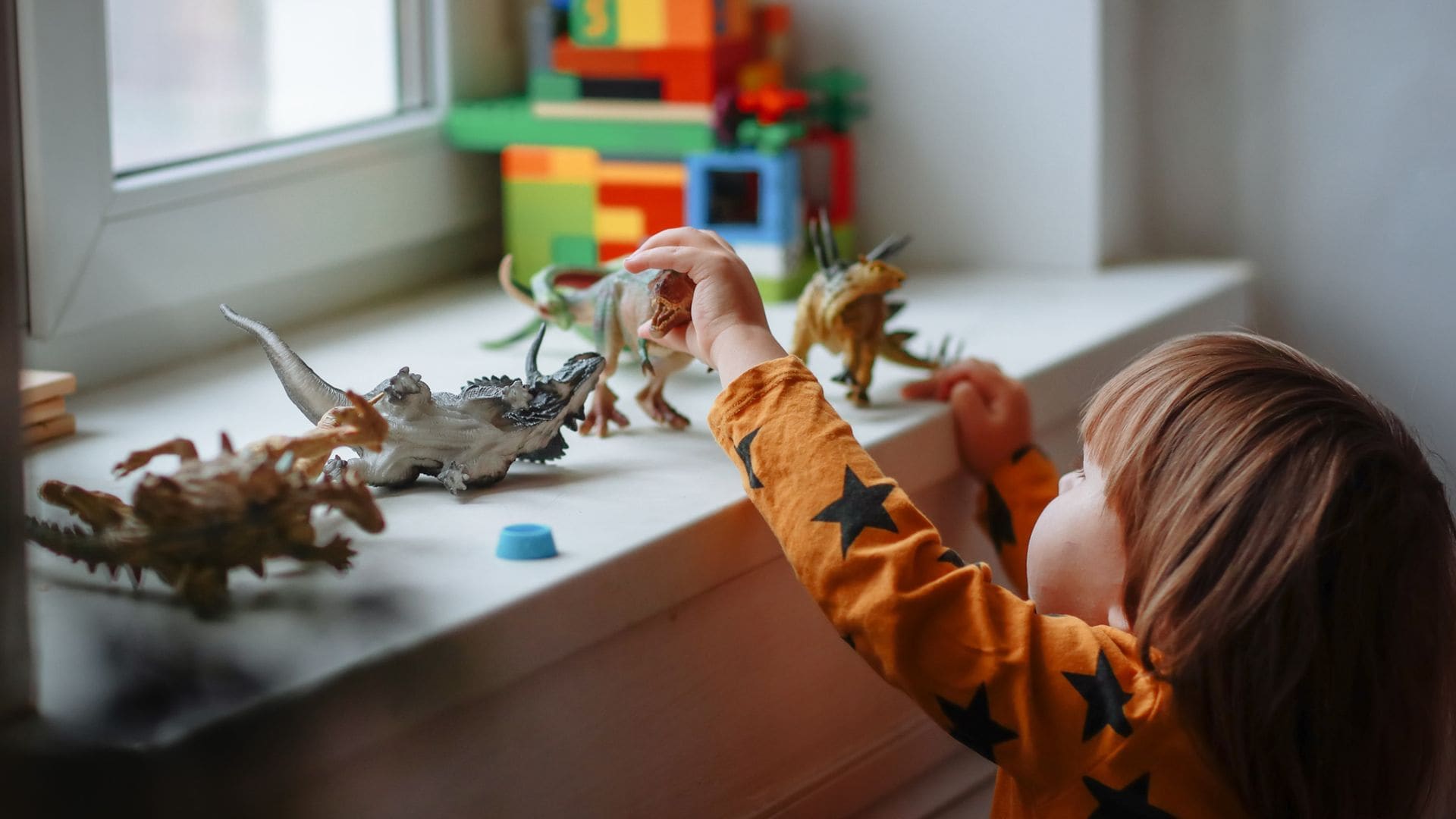 Niño pequeño jugando con dinosuarios de juguete en el alféizar de una ventana en blanco. El niño lleva una camiseta naranja con estrellas negras.