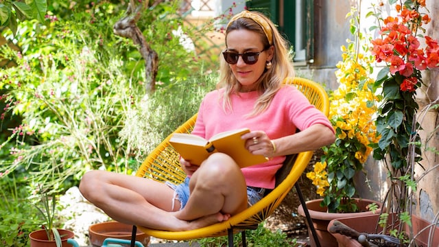 Mujer relajada leyendo en una silla de su jardín, rodeada de buganvillas y plantas, con un lazo en la cabeza, gafas de sol y ropa de primavera