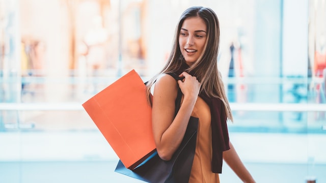 Chica joven comprando en un centro comercial, con bolsas al hombro y sonriendo mientras camina.