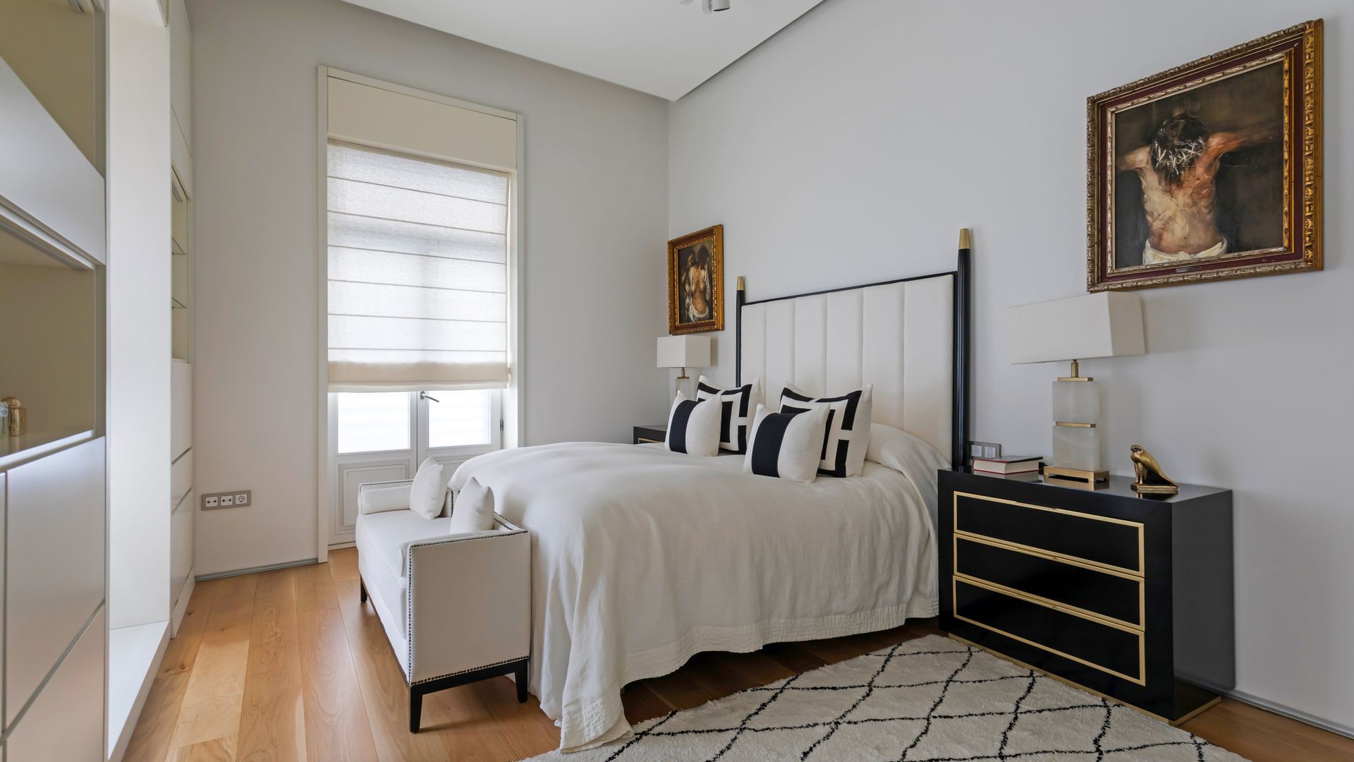 Dormitorio en blanco y negro con cama de hierro negra, mesita de noche negra con frente dorado, cuadro en la pared, banqueta a los pies de la cama, alfombra 