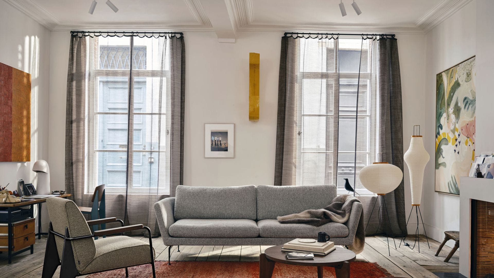 Salón con chimenea, alfombra marrón, molduras en el techo, sofá gris, lámparas de papel de pie, mesa auxilia, cuadros en las paredes, cortinas grises