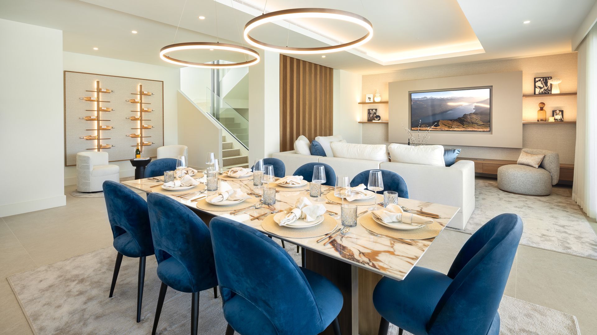 Salón comedor con comedor con sillas tapizadas en azul, mesa con el sobre de mármol, lámpara de techo, techo retroiluminado, estantería con luz con botellas de vino, televisión colgada en la pared