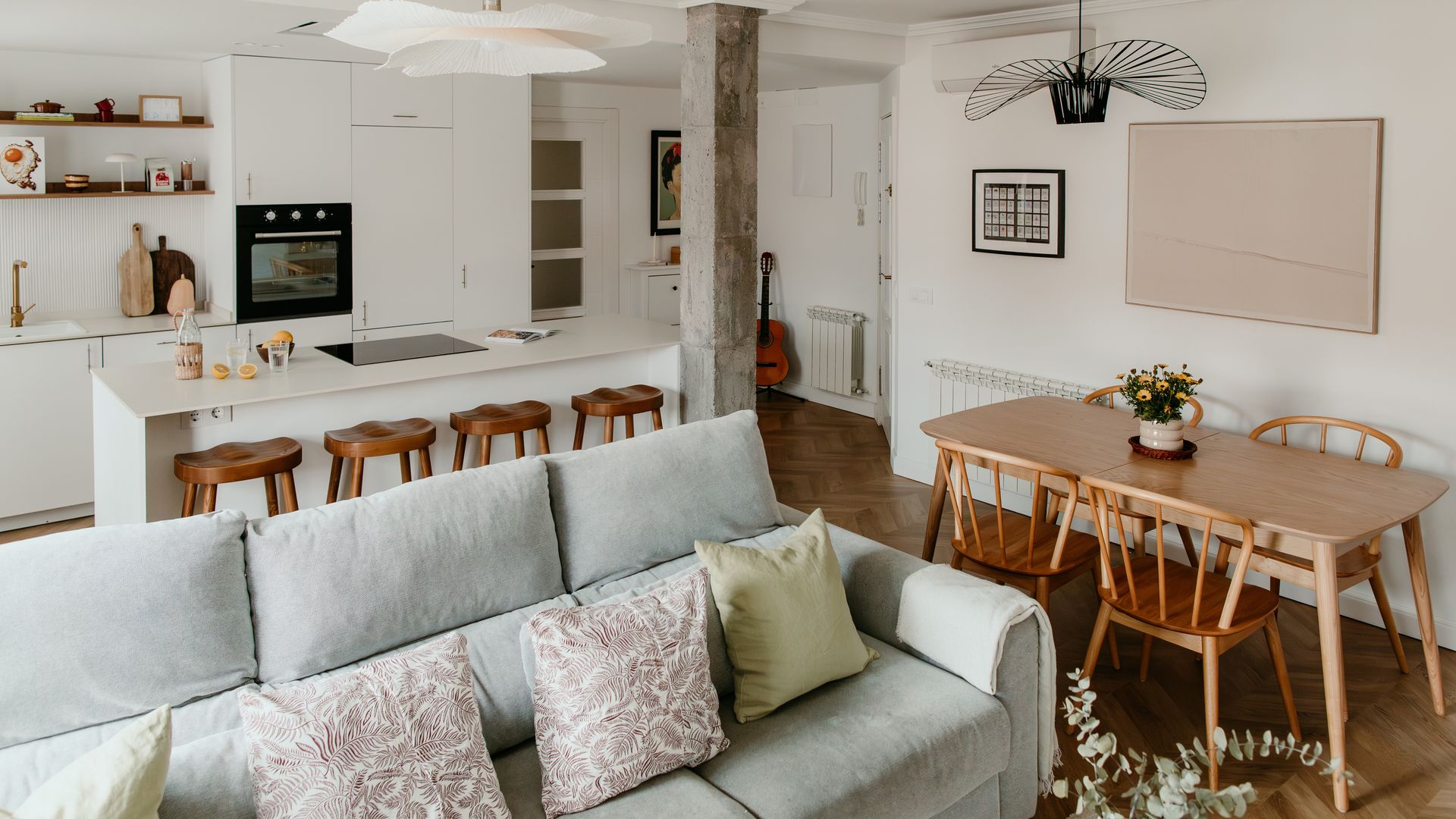Salón comedor y cocina, con columna de hormigón, sofá gris, cocina blanca, mesa de comedor de madera y sillas de madera, lámpara de techo negra