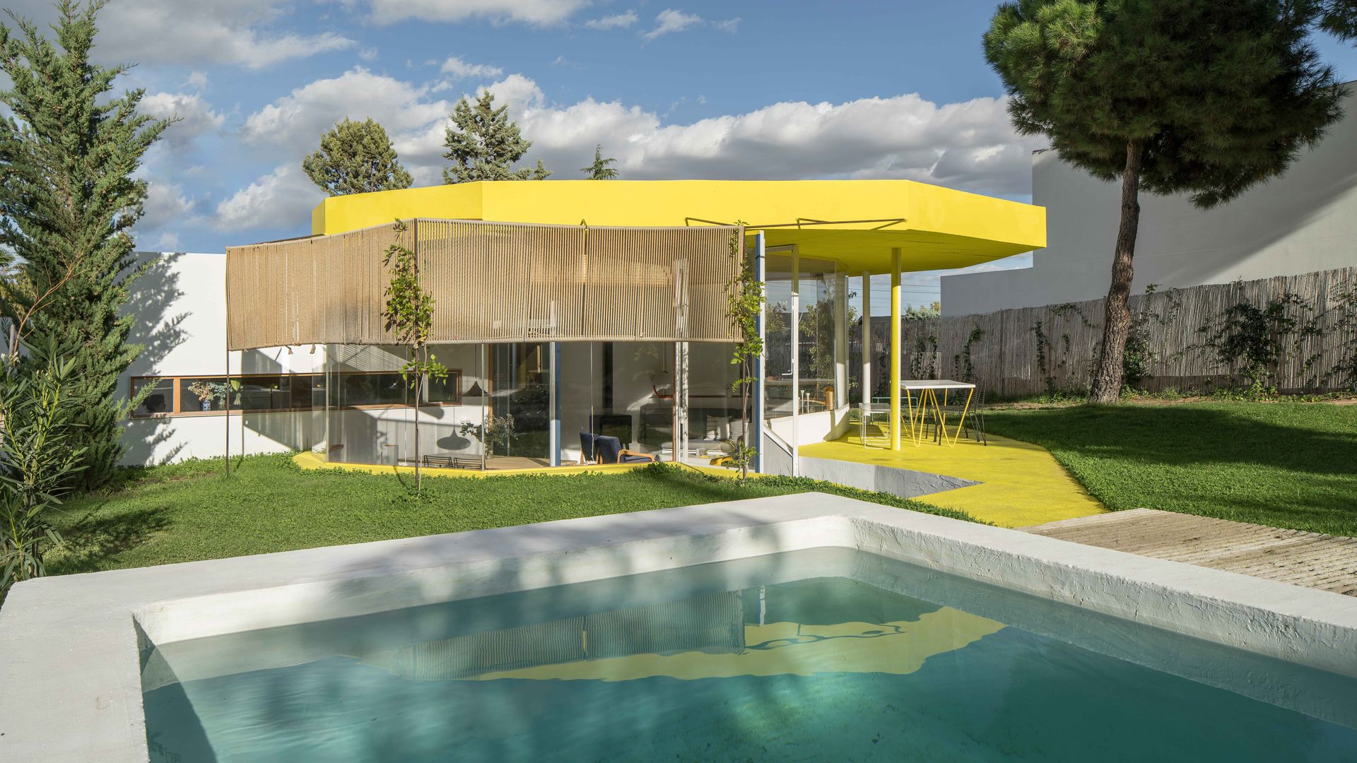 Casa con piscina, grandes ventanales y estructura amarilla, jardín con césped
