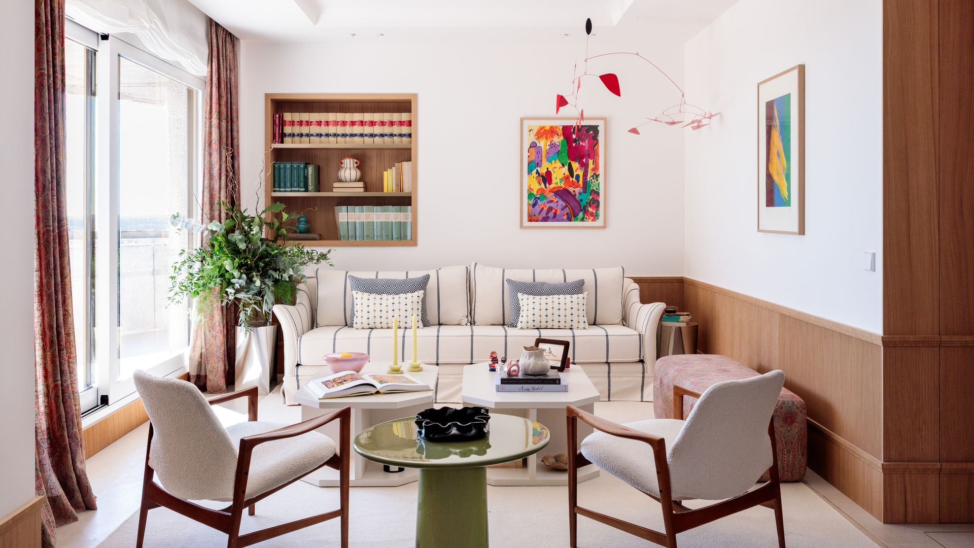 Salón con zócalo alto de madera, sofá blanco con rayas grises, cojines, cuadros de colores, rincón con dos butacas blancas y mesa auxiliar, mesas de centro, planta en el suelo, hornacina con estantería 