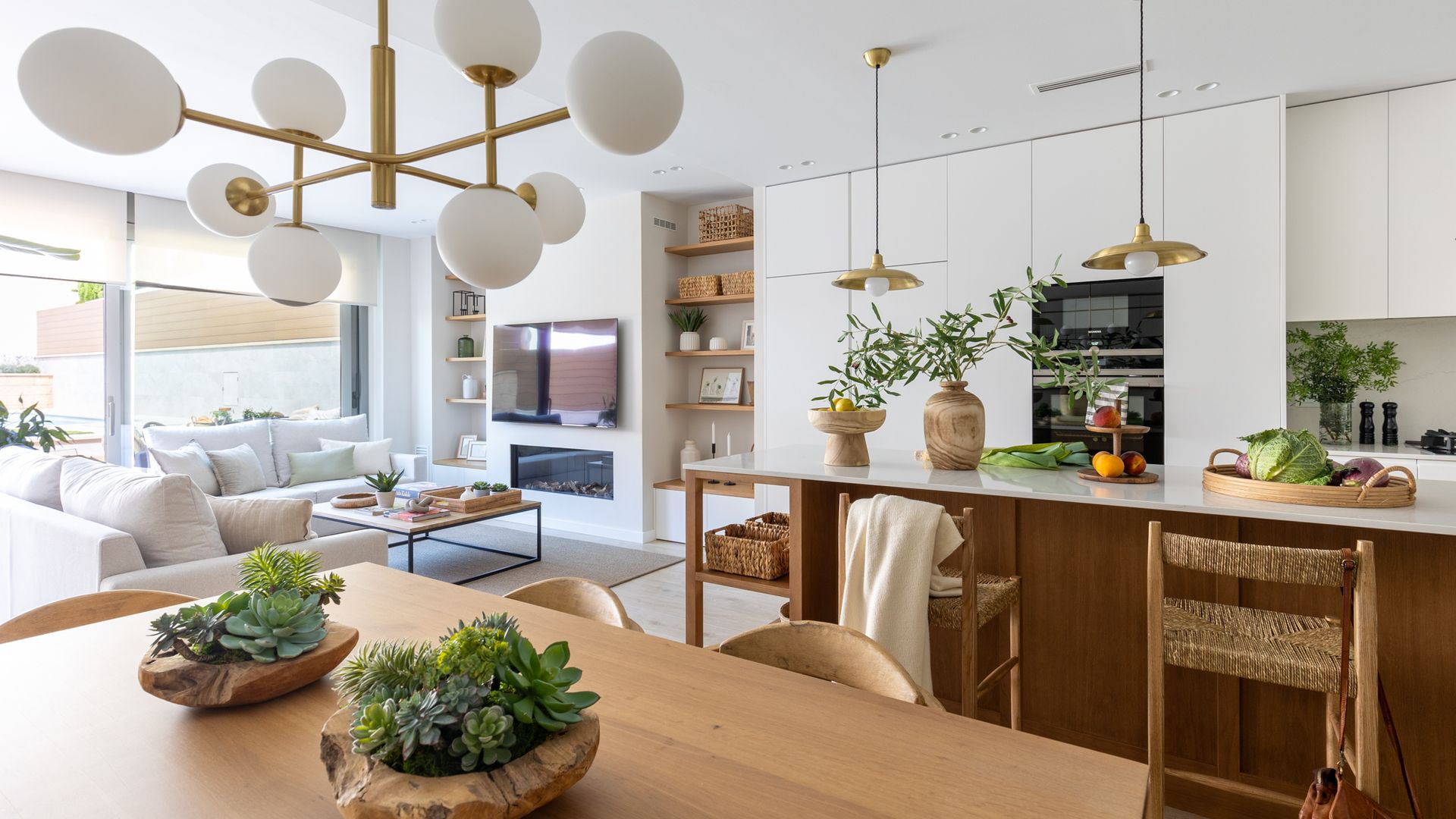 Salón comedor y cocina en blanco y madera con lámparas de techo, jarrones con plantas verdes 