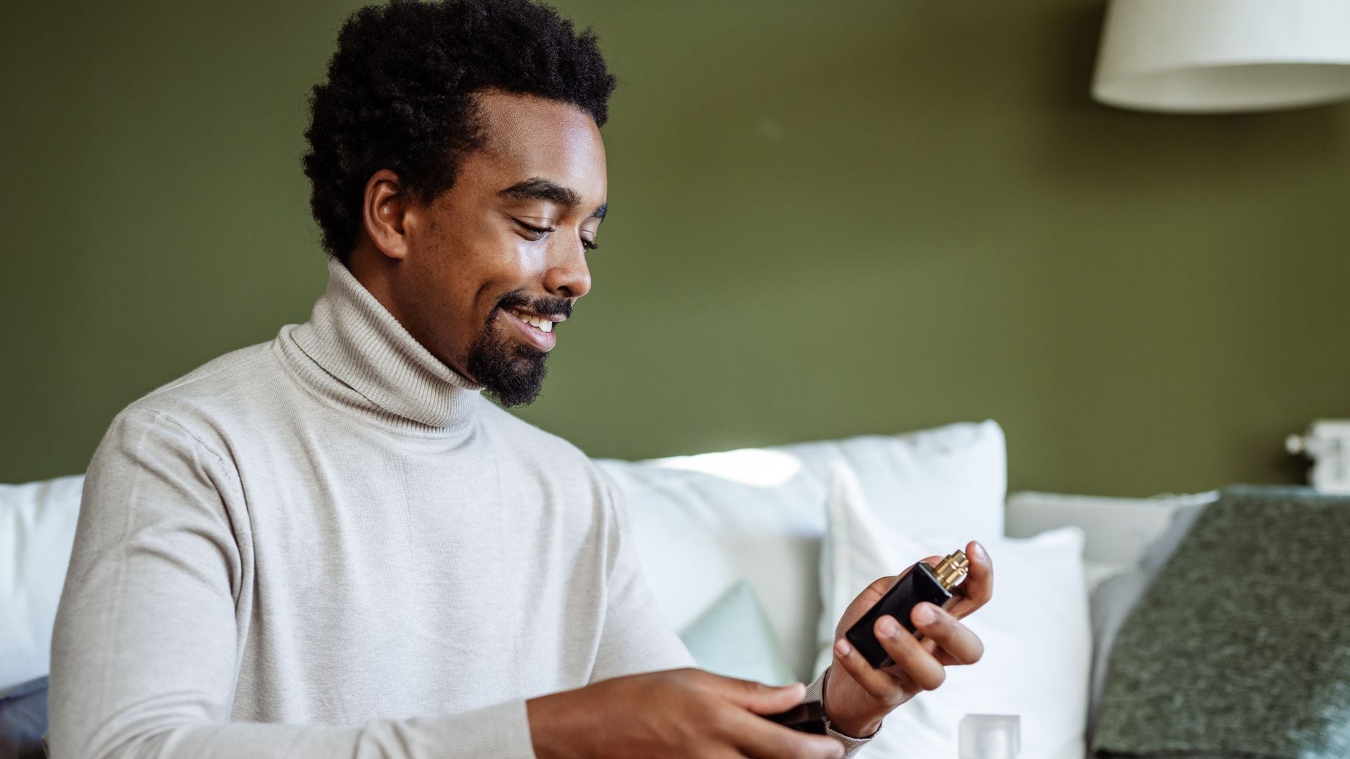 Hombre sentado en su sofá con jersey de cuello alto gris claro mirando perfumes