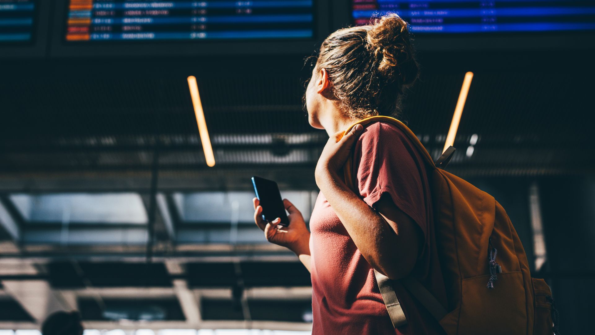 Mujer chequeando su vuelo en una pantalla del aeropuerto, con ropa de verano, teléfono móvil en la mano y mochila a la espalda.