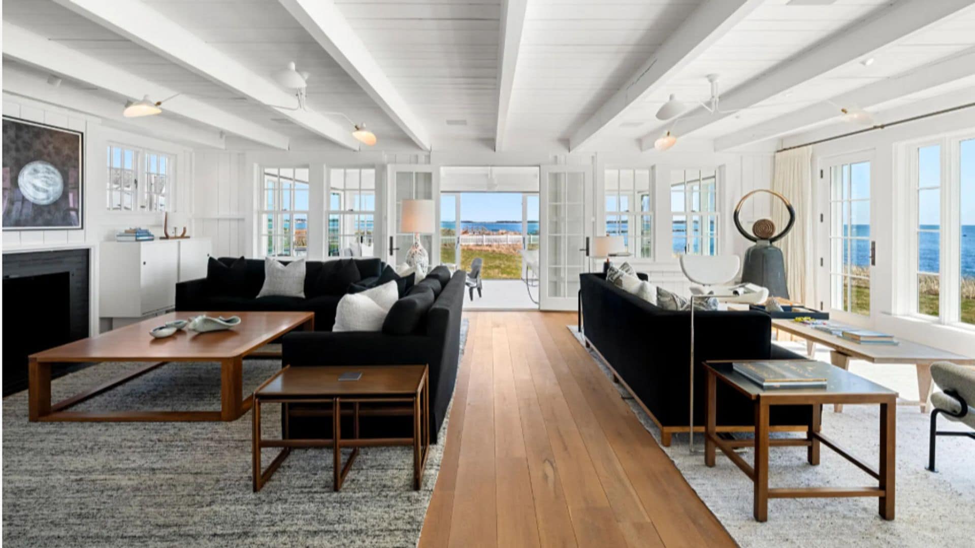 Salón abierto con dos zonas de estar, techos de madera blancos, suelo de madera, ventanales con cuarterones con vistas al mar, sofás negros, mesas de madera