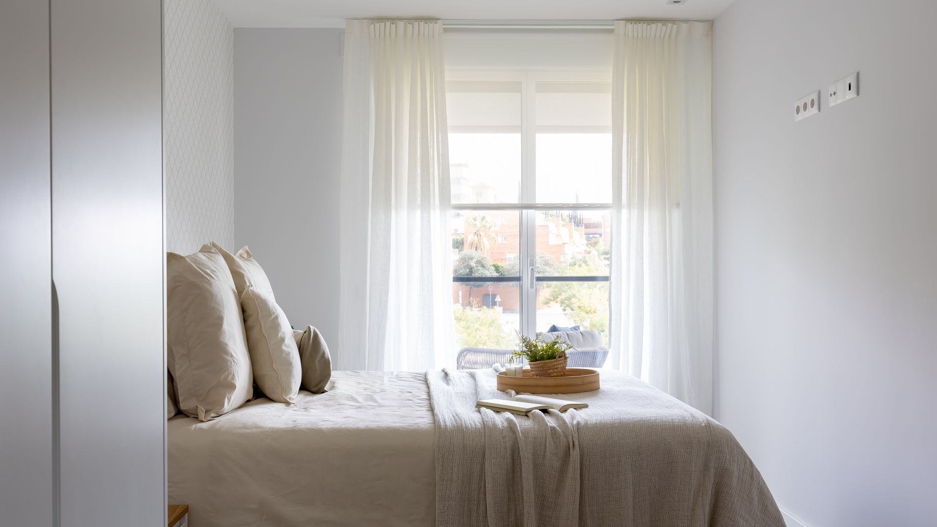 Dormitorio con cortinas blancas hasta el suelo, ropa de cama en tonos beige, cojines, bandeja de madera sobre la cama, papel pintado en la pared del cabecero 