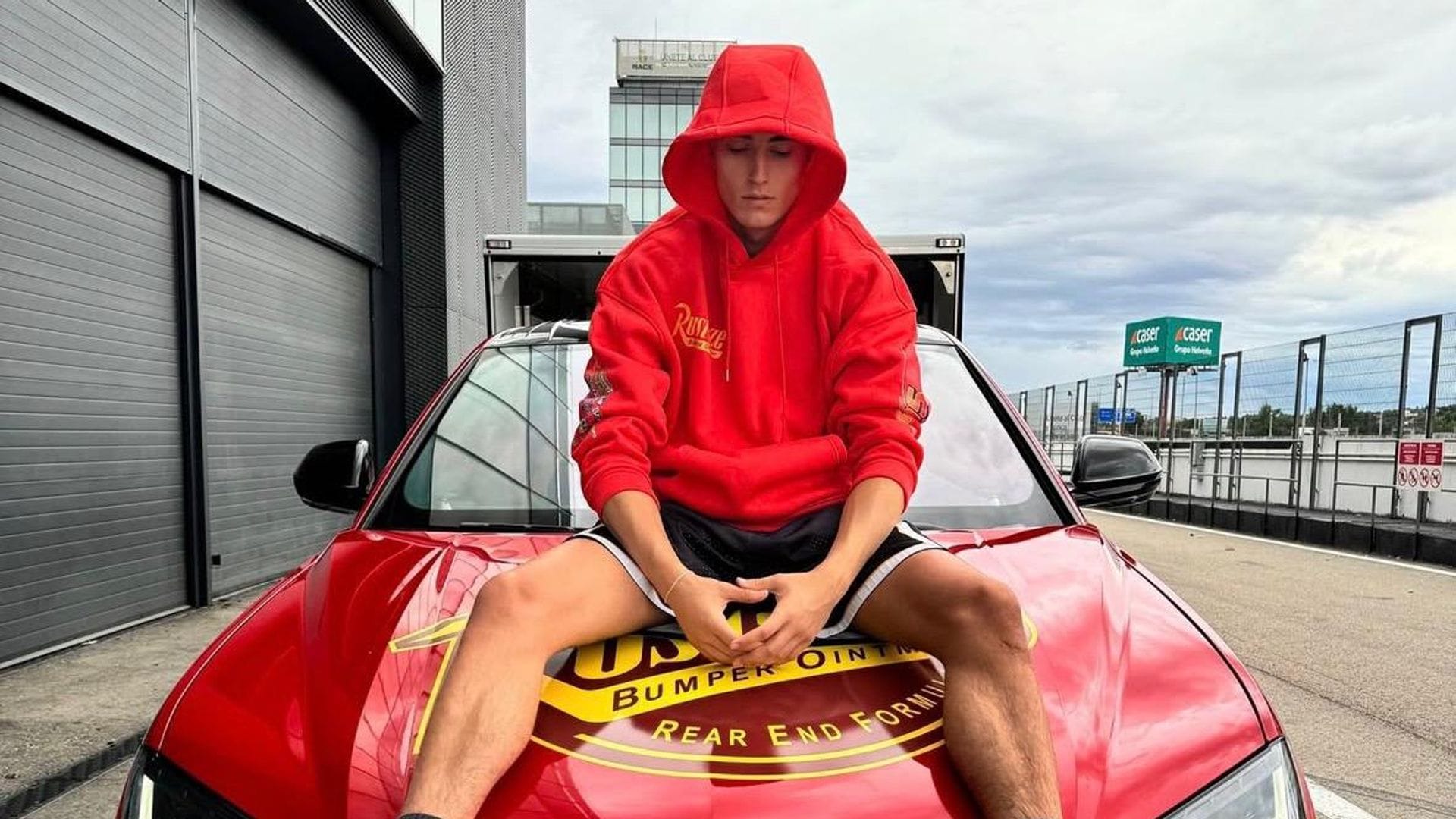 Plex en sudadera roja y zapatillas, sentado en un Lamborghini rojo.