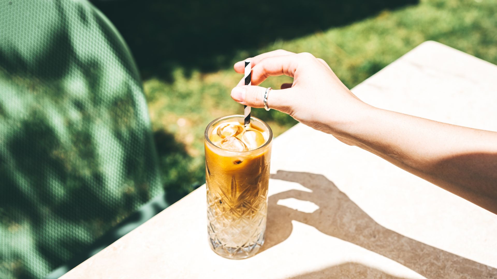 Café con hielo en vaso de cristal, con la mano de una chica removiéndolo con una pajita apoyado sobre la mesa de un jardín.