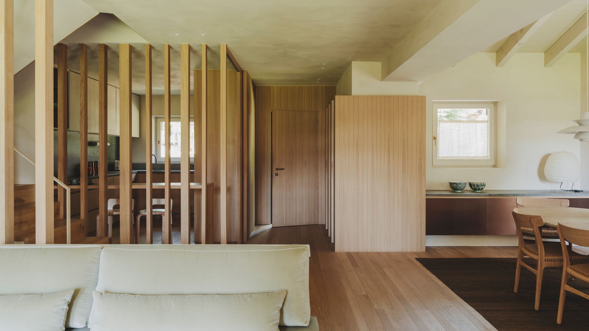 Salón comedor abierto a la cocina y a la puerta de entrada, con suelo y tabiques de madera, estructura de lamas independizando la cocina, sofá blanco, mesa de comedor de madera