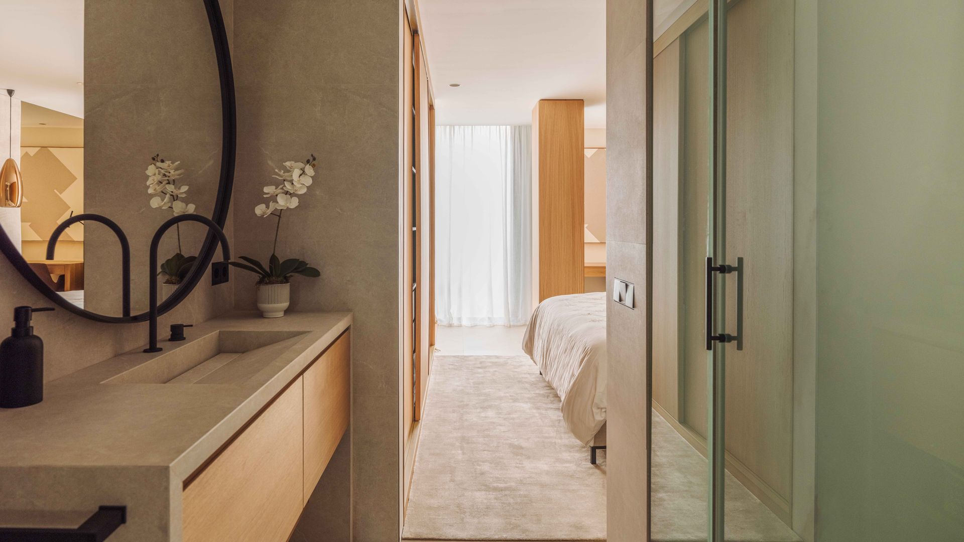 Dormitorio con baño integrado con ducha, mampara de cristal traslucido, espejo redondo con el marco negro, grifería negra y mueble volado de madera 