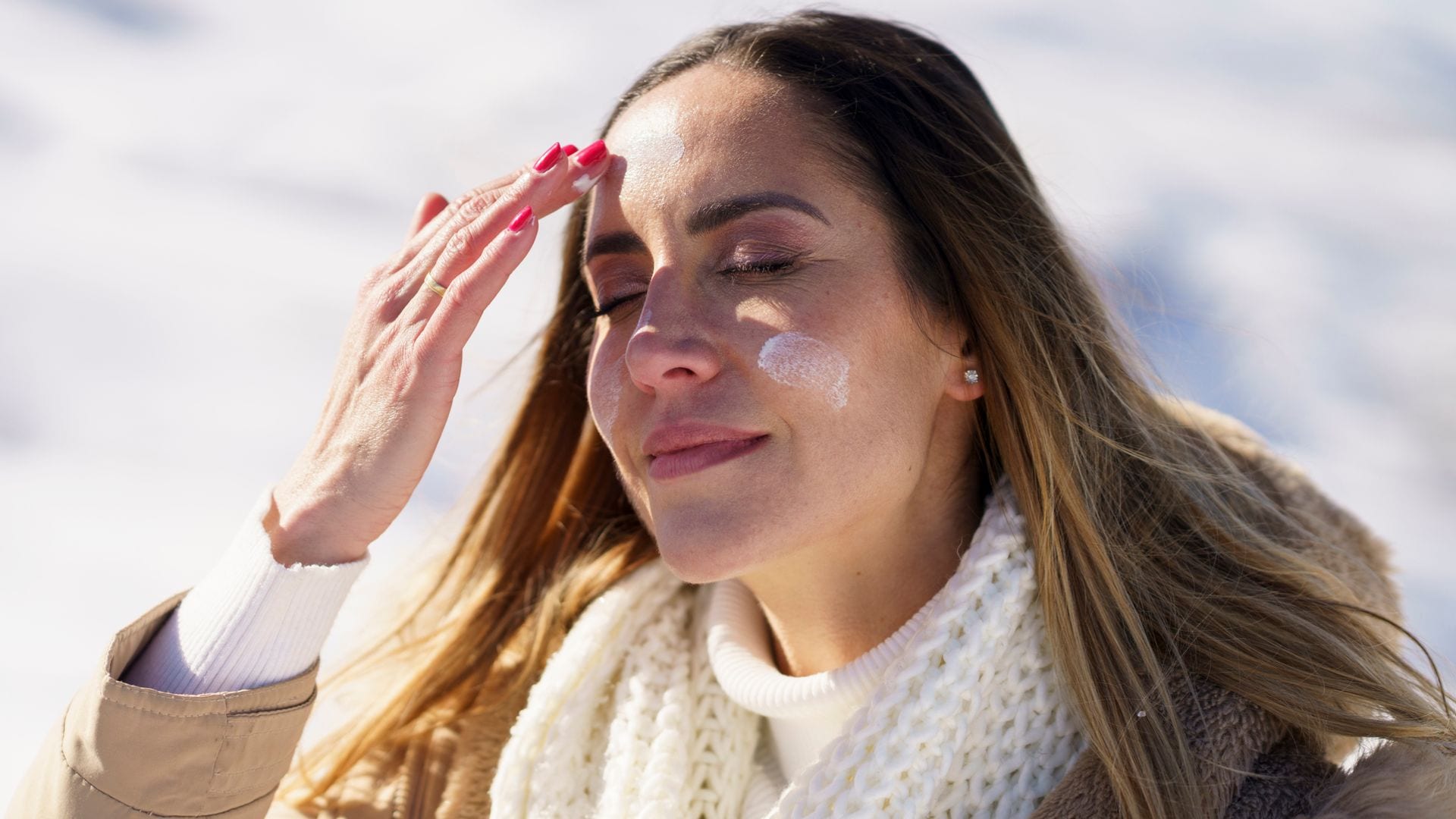 Mujer en la nieve aplicándose crema en el rostro, con abrigo beis y bufanda blanca