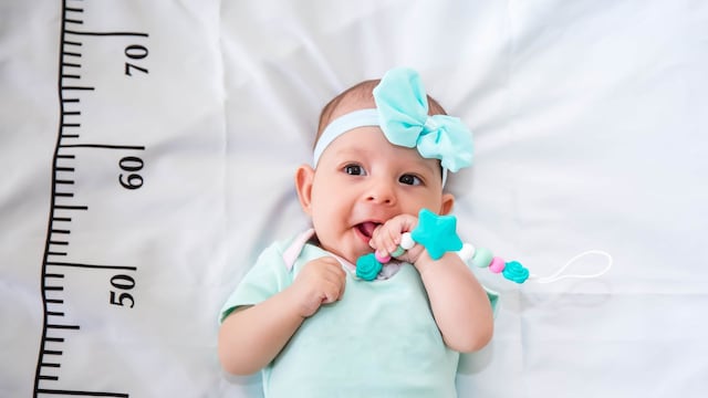 Un bebé de tres meses, vestido de verde menta, acostado en una cama sobre la que está dibujado un medidor de crecimiento. Con un mordedor en las manos, masticando.