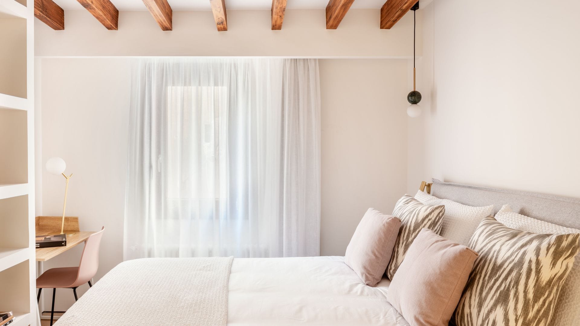 Dormitorio con vigas de madera, escritorio, estantería, cabecero tapizado, cortinas blancas, lámpara de techo tipo bombilla