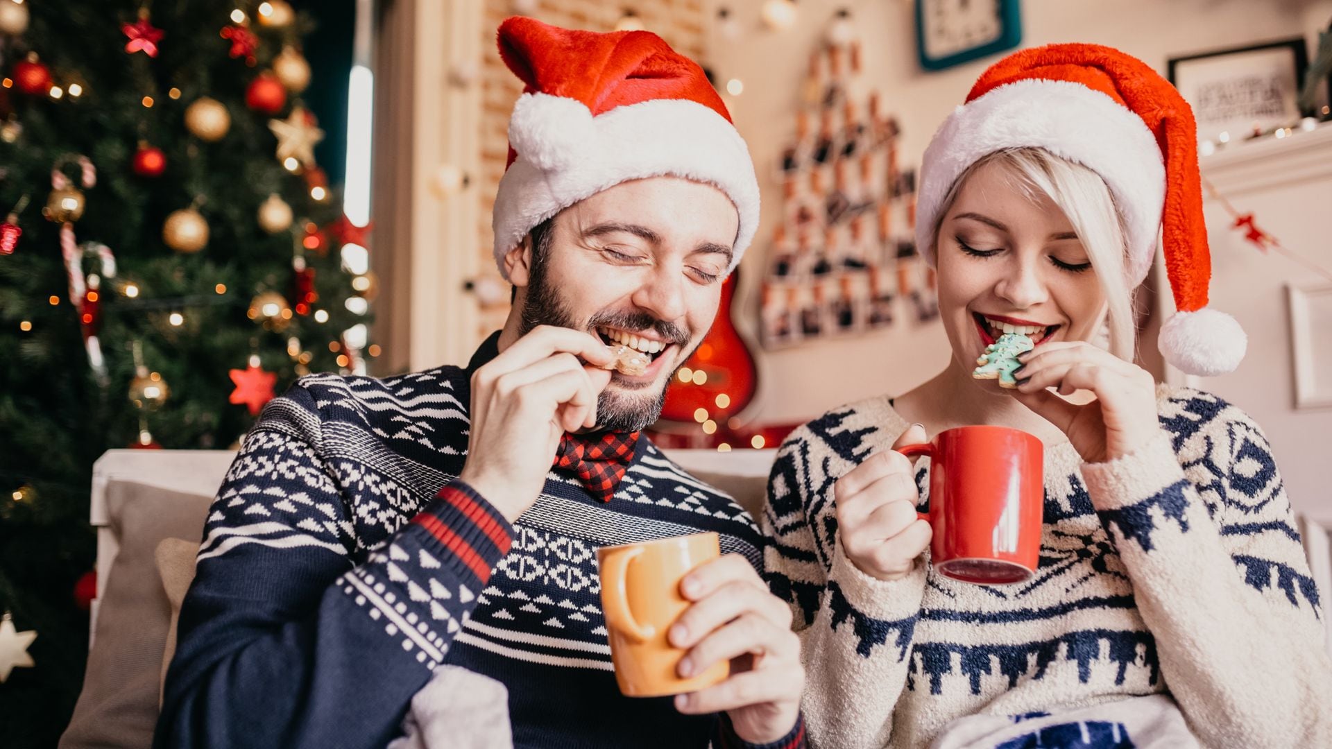 pareja con gorros de Papá Noél comiendo galletas y chocolate caliente en casa, en Navidad