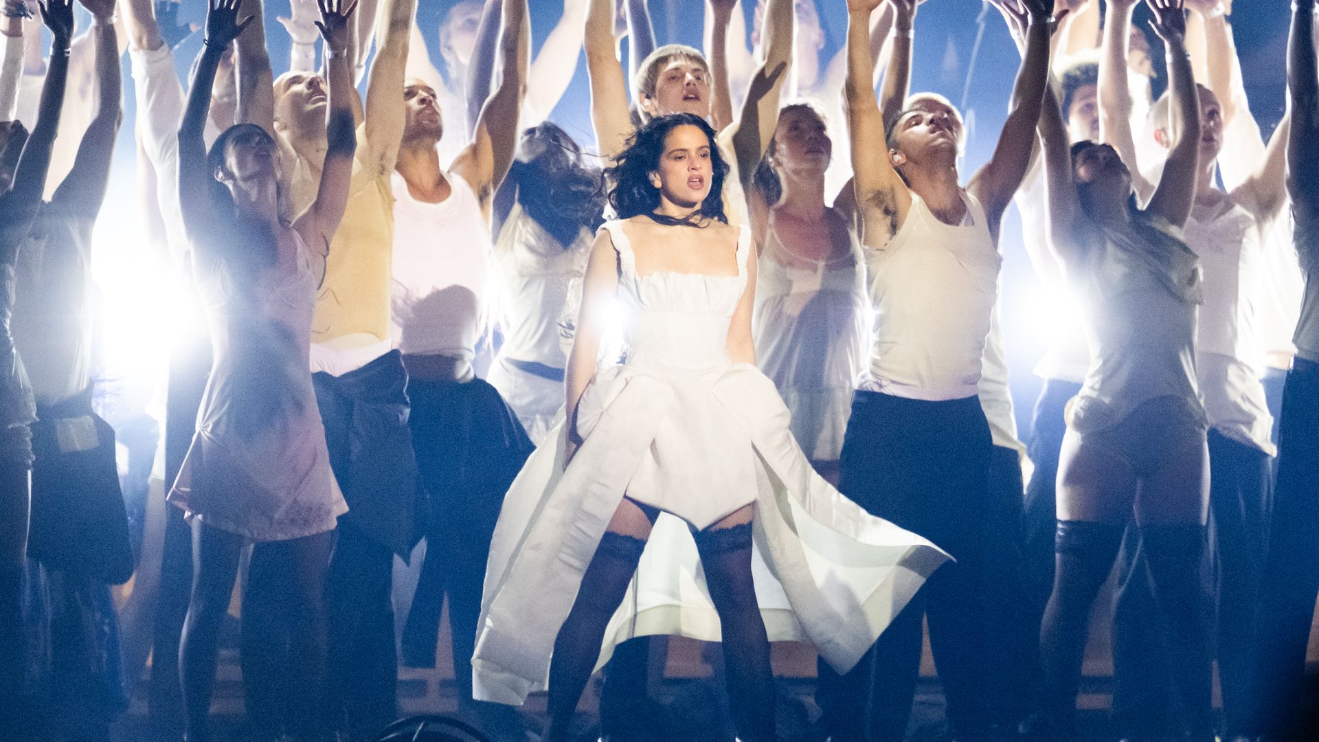 Rosalía, en un vestido blanco, actúa en un escenario con un coro elevando las manos.