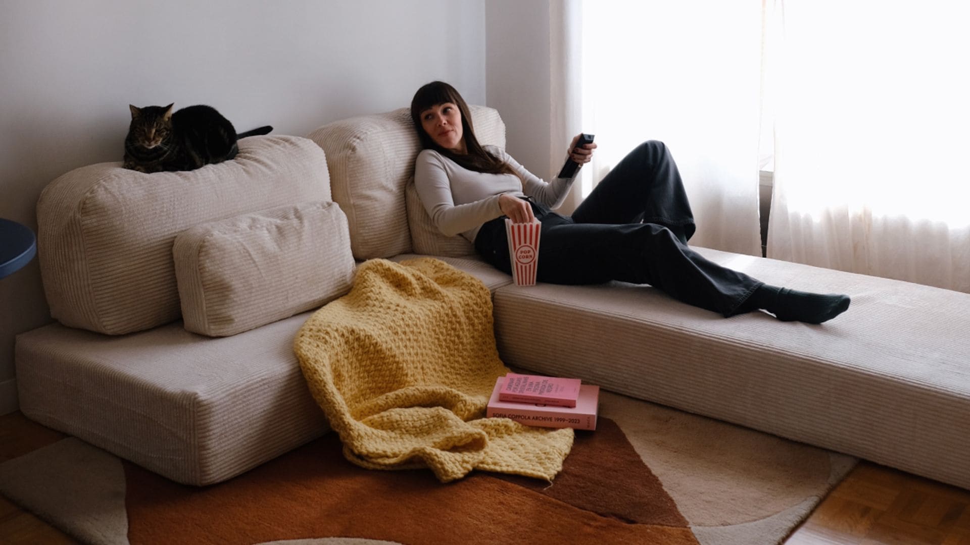 Mujer tumbada en un sofá modular de color beige y en pana, alfombra marrón, gato en el respaldo, visillos blancos