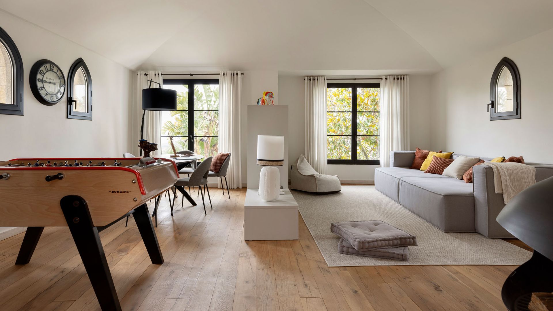 Salón comedor con futbolín, sofá gris, mesa de comedor, alfombra, cojines de suelo, mueble bajo separando el salón del comedor, suelo de madera