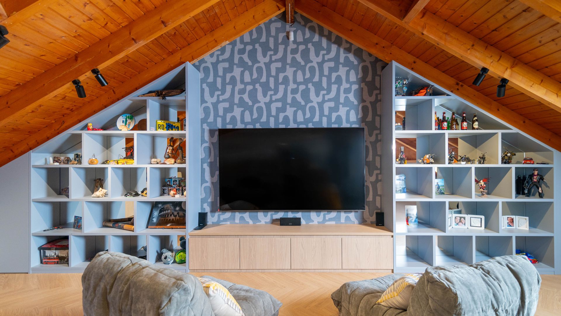 Buhardilla con el techo de madera abuhardillado, mueble de televisión, televisión colgada en la pared, papel pintado y estanterías azul claro a los lados