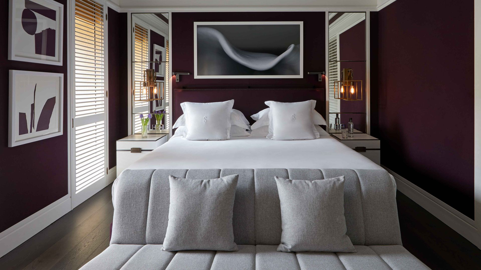 Dormitorio con paredes en color burdeos, sábanas blancas, banco tapizado en gris a los pies de la cama, cuadro en la pared del cabecero, molduras techo 