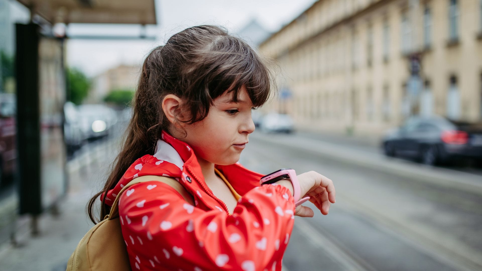 Niña en la calle con una mochila y un Smartwatch en la muñeca hablando con él. Lleva ropa de otoño-invierno porque acaba de llover.