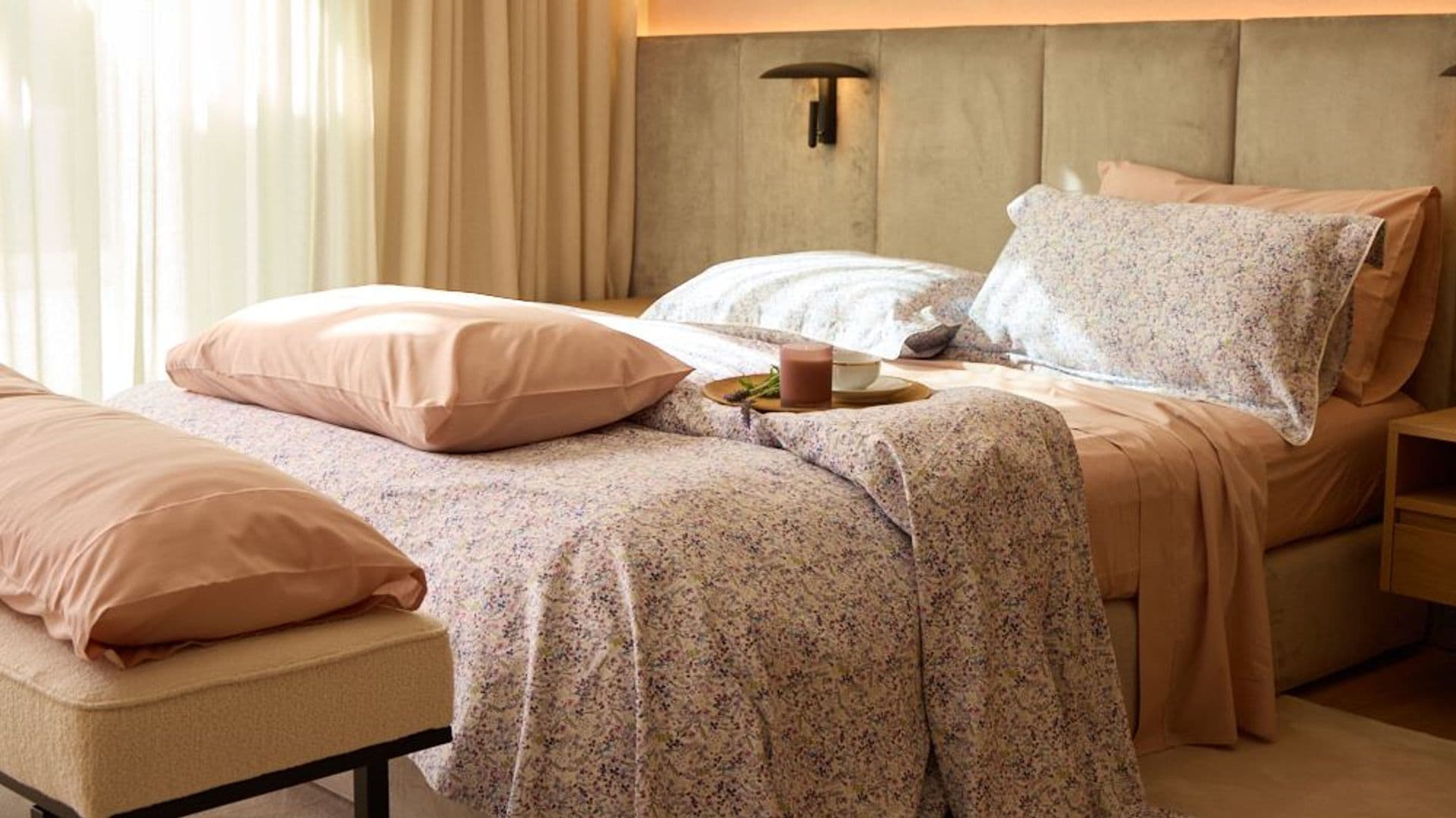 Dormitorio con cortinas beiges, cama con cabecero tapizado en verde, funda nórdica de flores, aplique en el cabecero, banqueta a los pies de la cama, almohadas y bandeja con desayuno sobre la cama