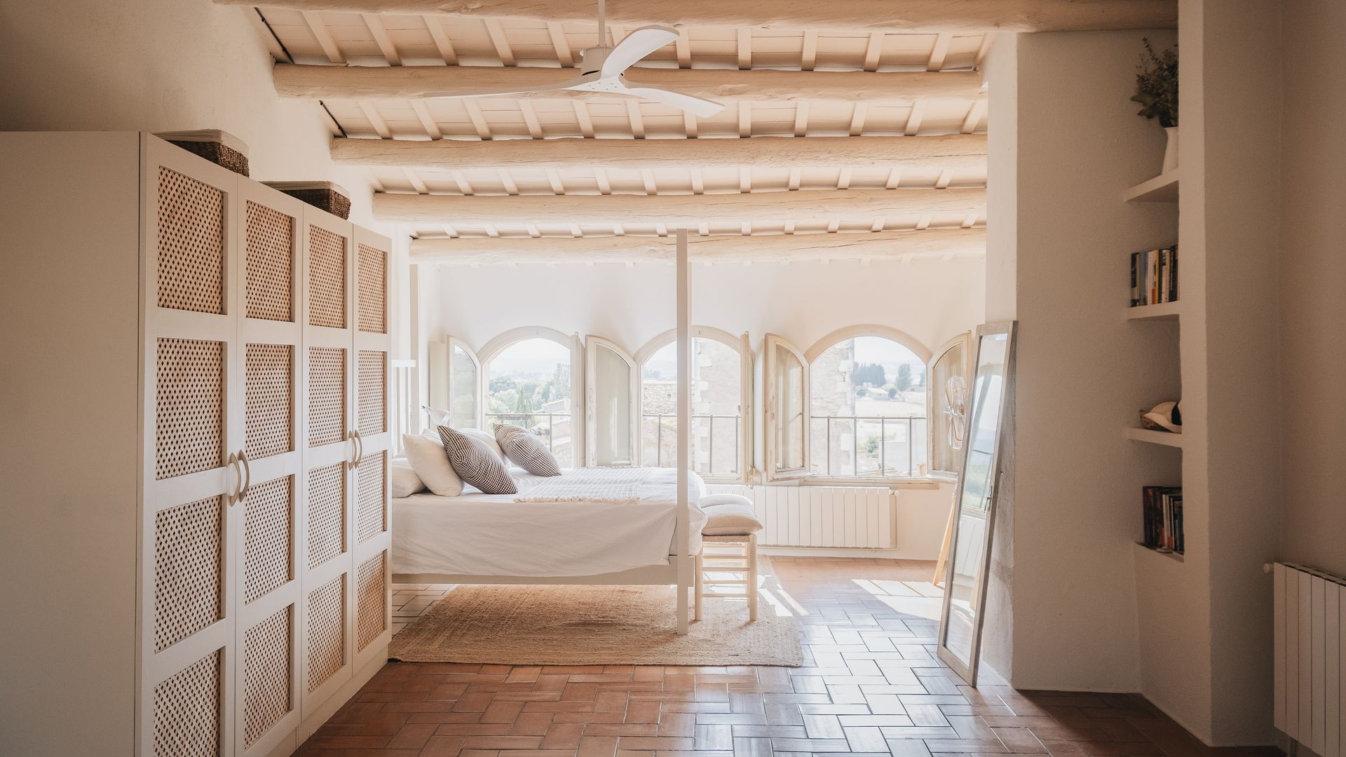 Dormitorio con armario exento con puerta de rejilla, librería de obra, suelo de barro, ventanas en forma de arco, ventilador de techo, banqueta, cama con dosel