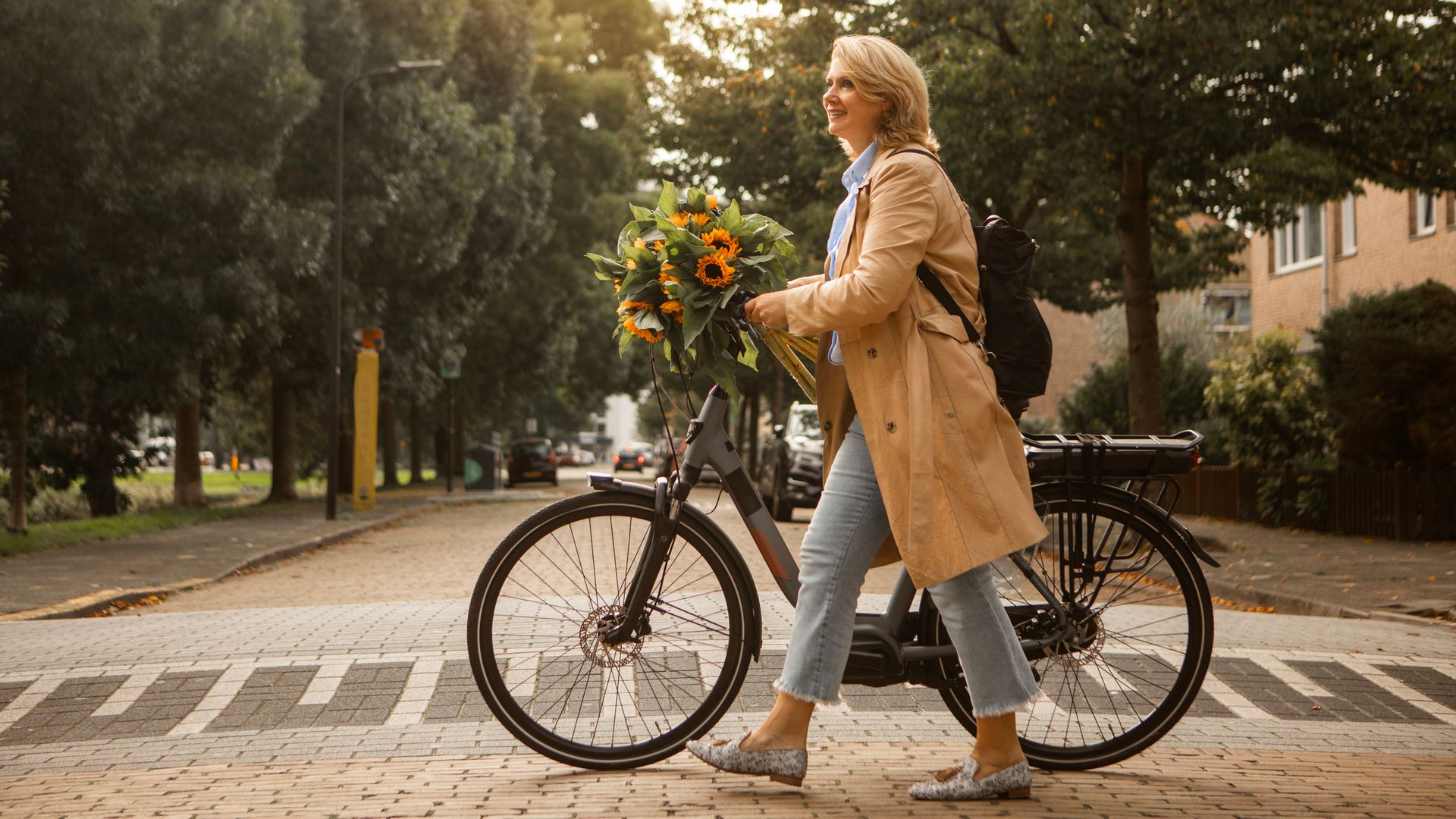 Mujer adulta de 50 años y rubia paseando con su bicicleta por la calle, cruzando, con flores en la mano mientras sujeta el manillar. Lleva ropa de otoño o primavera, con gabardina beige, pantalones jeans capri y bailarinas.