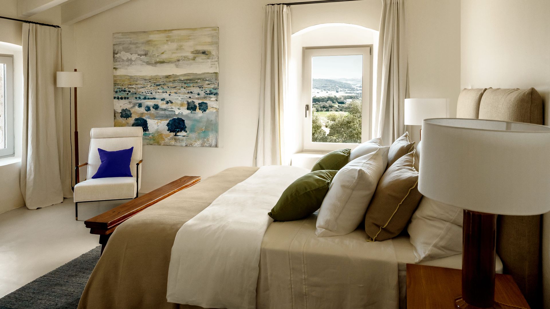 Dormitorio con cama con cojines, almohada, plaid, rincón lectura con sillón blanco, lámpara de pie, cortinas