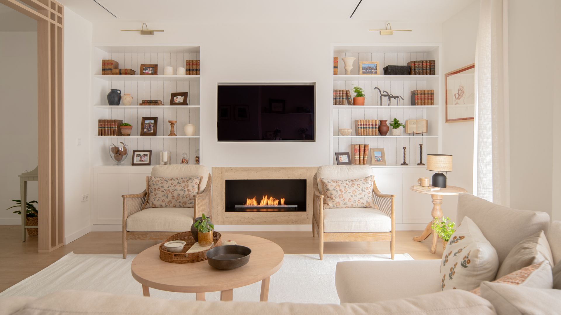 Salón con chimenea encendida, mueble a medida con estanterías a los lados, blancos, con trasera de lamas blancas, sofá beige, alfombra, butacas de madera tapizadas, mesa de centro de madera
